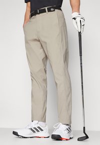 Pantalons de golf beiges avec poches latérales, une ceinture tressée noire, associés à des chaussures de golf blanches. Le golfeur tient un club dans la main droite.