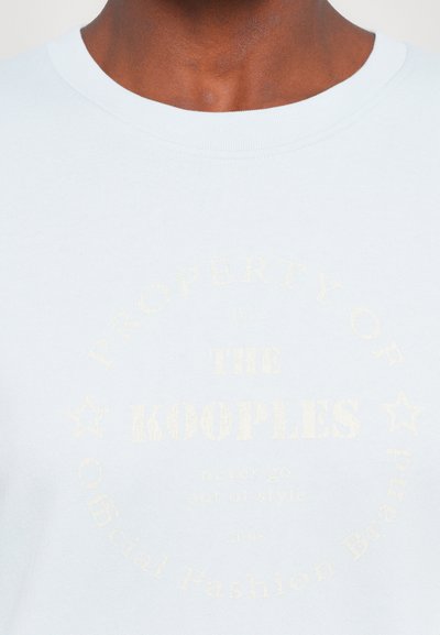 Lyseblå t-skjorte med korte ermer laget av myk bomull, med et sirkulært logo i lys gulltekst trykket foran.