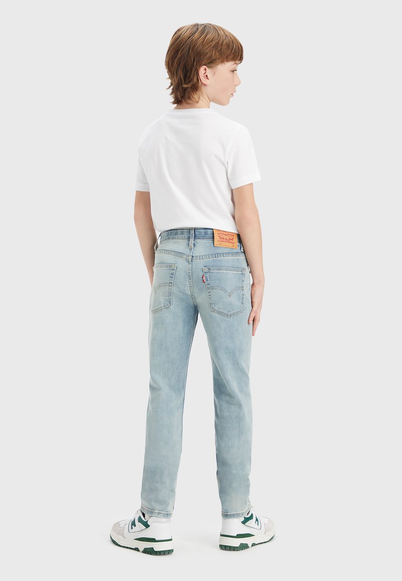 Vaqueros de mezclilla azul claro con un diseño de pierna recta, bolsillos traseros y una etiqueta de marca de cuero marrón en la cinturilla. Camiseta blanca y zapatillas.