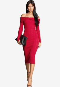 Abito rosso a coste, off-the-shoulder, con maniche lunghe e dettaglio a fiocco, silhouette aderente, lunghezza al ginocchio, abbinato a una pochette nera.