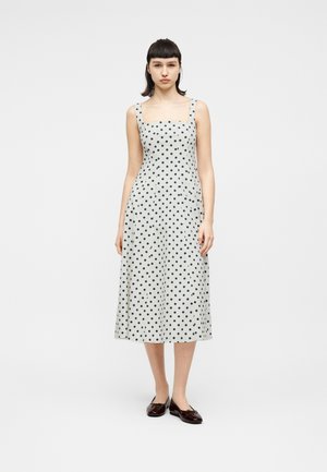 Frau steht, trägt ein weißes ärmelloses Midikleid mit schwarzen Punkten und dunkle flache Schuhe, vor einem schlichten hellen Hintergrund.