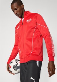 Red athletic zip jacket with white accents and logo. Soft fabric, fitted design, and patterned sleeve details. Black pants with logo.