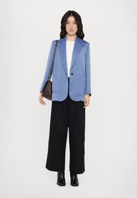 Lyseblå blazer med en enkelt svart knapp og to frontlommer, båret over en hvit topp og kombinert med svarte vide bukser og svarte støvler.