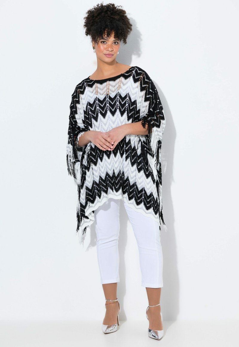 Svart och vit poncho med zigzag-mönster och fransade kanter, tillverkad av ett lätt material, bärs över vita tajta byxor och silvriga klackar.