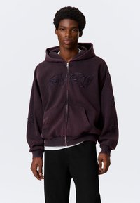 Paarse hoodie met ritssluiting en capuchon, met geborduurde tekst en sterpatronen op de mouwen. Gemaakt van een zacht, gestructureerd materiaal.