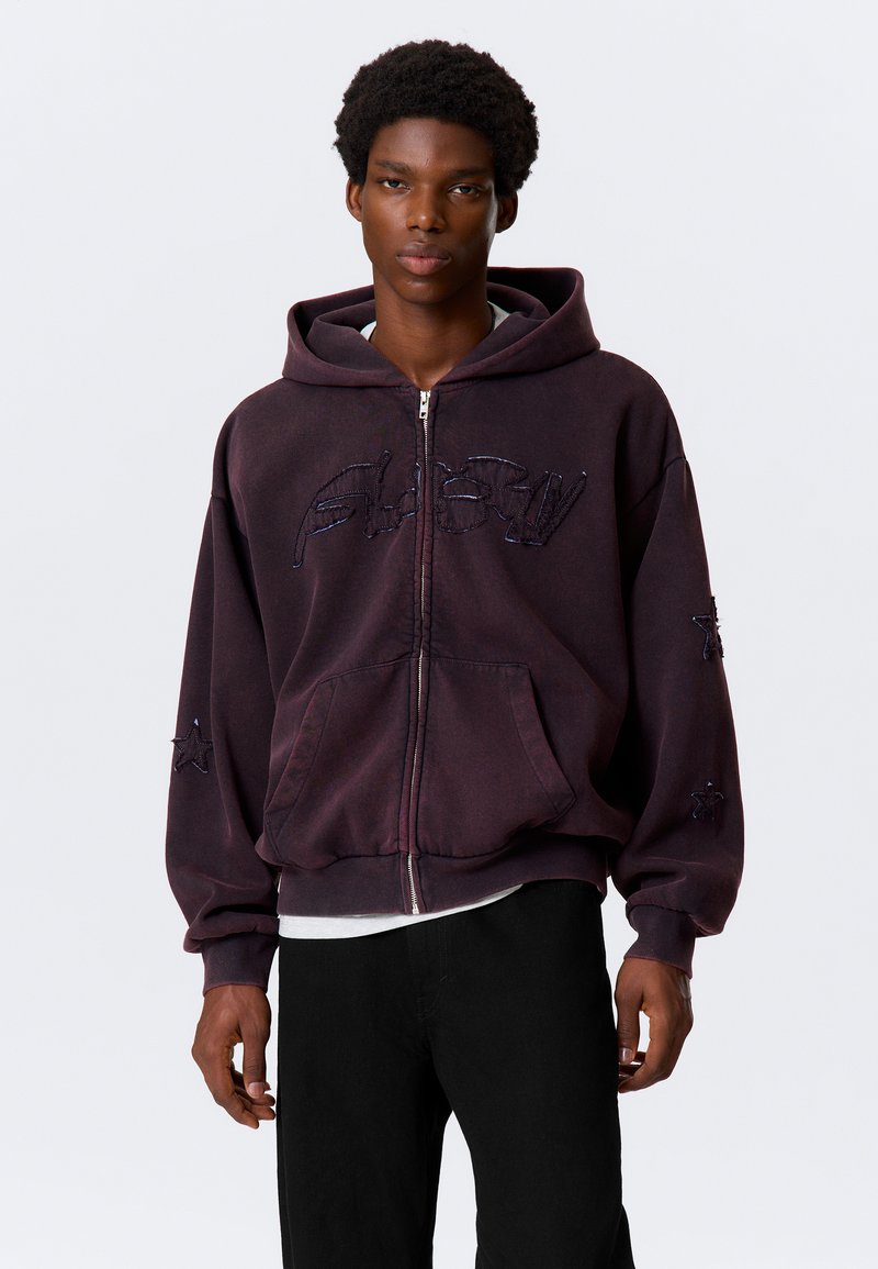 Paarse hoodie met ritssluiting en capuchon, met geborduurde tekst en sterpatronen op de mouwen. Gemaakt van een zacht, gestructureerd materiaal.