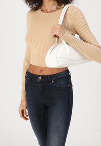 Weiße Lederhandtasche mit abgerundeter Form und glatter Textur, getragen von einer Person in einem beige gerippten Oberteil und dunklen Skinny-Jeans.