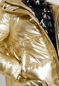 Gouden pufferjack met een glanzende, reflecterende textuur. Voorzien van ritssluiting, een capuchon en zakken. De binnenkant heeft een zwart grafisch patroon.