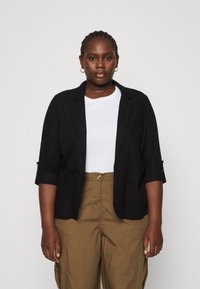 Blazer nero con una vestibilità rilassata, maniche arrotolate e due tasche, indossato su una camicia bianca e abbinato a pantaloni verde oliva.