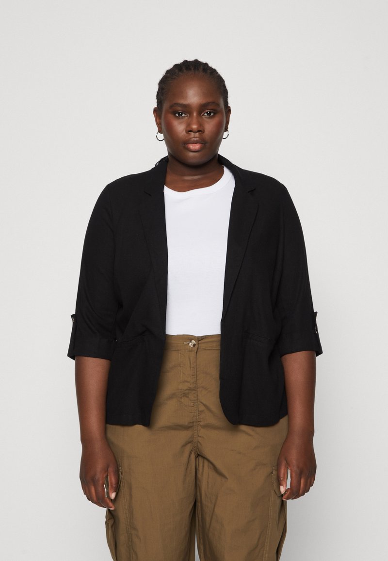Blazer nero con una vestibilità rilassata, maniche arrotolate e due tasche, indossato su una camicia bianca e abbinato a pantaloni verde oliva.