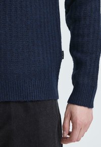 Navy knitted sweater featuring a ribbed texture with a raised pattern. The hem and cuffs are ribbed, with a small logo tag on the side.