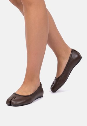 Jambes portant des ballerines en cuir marron foncé à bout fendu sur un fond clair uni, un pied légèrement levé.