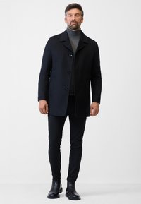 Zwarte wolmix jas met een kraag, vier knopen en zijzakken. Draagt boven een grijze coltrui en gecombineerd met smalle zwarte broek en laarzen.