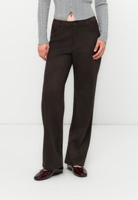 Mujer vestida con pantalones rectos marrón oscuro, suéter de punto trenzado gris y bailarinas brillantes de color rojo oscuro, de pie frente a un fondo blanco.