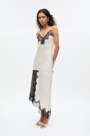 Femme portant une robe nuisette crème sans manches avec un bord en dentelle noire et des talons ouverts beige, debout devant un fond blanc uni.