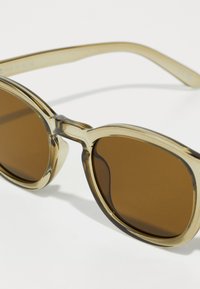 Lunettes de soleil teintées de marron avec une monture transparente de couleur marron clair. Les verres ronds et les branches épaisses présentent une texture lisse et brillante.