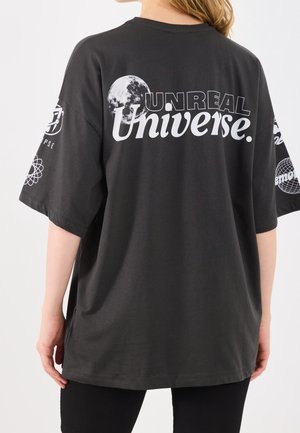 Person trägt ein schwarzes übergroßes T-Shirt mit weißem Schriftzug "UNREAL Universe" und einer Mondgrafik auf der Rückseite sowie wissenschaftlich inspirierte Logos auf den Ärmeln.