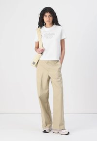 T-shirt blanc court avec imprimé "Carhartt WIP", pantalon beige à jambes larges et sac à bandoulière crème ; le mannequin porte des baskets claires.