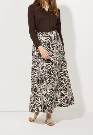 Vrouw met een zwart shirt met lange mouwen, een lange rok met zwart-witte zebraprint en beige instapsandalen, staand tegen een witte muur.