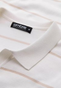 Valkoinen poolopaita, jossa on vaaleanbeigejä vaakasuoria raitoja, valmistettu teksturoidusta kankaasta. Varustettu tavallisella kauluksella ja mustalla tuotemerkki-etiketillä sisällä.