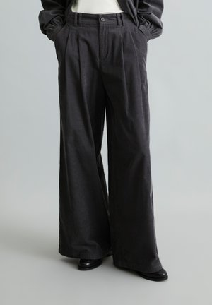 Personne portant un pantalon noir taille haute à jambes larges, les mains dans les poches et des chaussures noires, debout devant un fond gris uni.