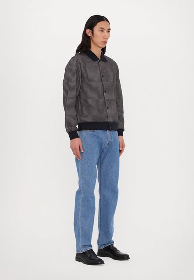 Μπουφάν bomber με κουμπιά σε υφασμάτινο navy και μπεζ σχέδιο, με μαύρο γιακά και ριμπ μανσέτες, σε συνδυασμό με ελαφριά μπλε τζιν με ίσια γραμμή.