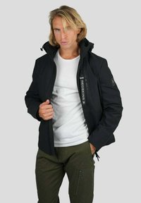 Veste noire imperméable avec fermeture éclair, col haut et détails de logo. Associée à un t-shirt blanc et un pantalon cargo vert. Fond uni.