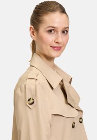 Vaaleanbeige trenssi, jossa on leveä kaulus, kaksirivinen nappisulku ja olkapäät; sileä kangas perinteisellä muotoilulla.