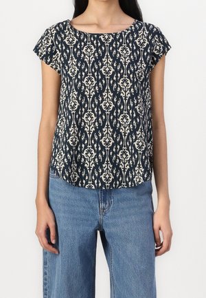 Femme portant un haut bleu marine à manches courtes avec un motif blanc complexe et un jean bleu taille haute sur fond blanc.