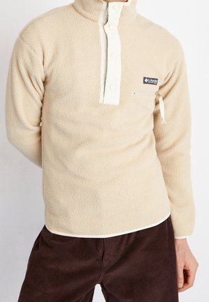 Beige fleece trui met witte drukknoppen en een borstzak met rits, gedragen met donkerbruine corduroy broek.