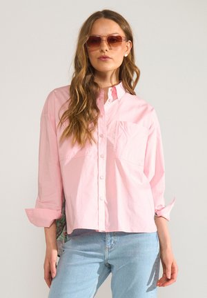 Vrouw met lang golvend haar, die een oversized lichtroze blouse met knopen draagt, lichtblauwe spijkerbroek, roze zonnebril en pareloorbellen tegen een effen achtergrond.