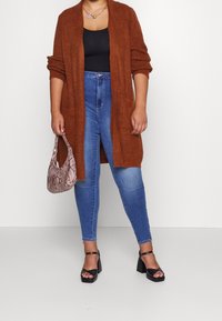 Cardigan lavorato a maglia color ruggine con texture a coste, indossato sopra una maglietta nera, abbinato a jeans blu a vita alta e sandali neri con tacco bloccato.