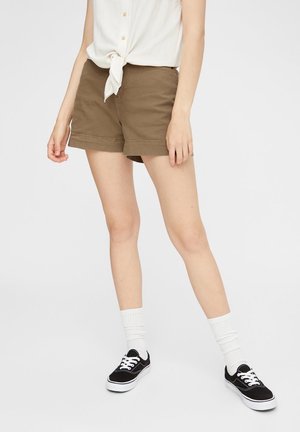 Personne portant un short marron, une chemise blanche nouée, des chaussettes blanches et des baskets noires, debout devant un fond clair uni.