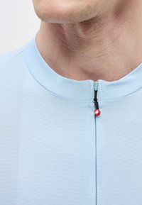 Mand iført lyseblå skjorte med kort krave og lynlås foran med en lille rød trækflig med hvidt logo.