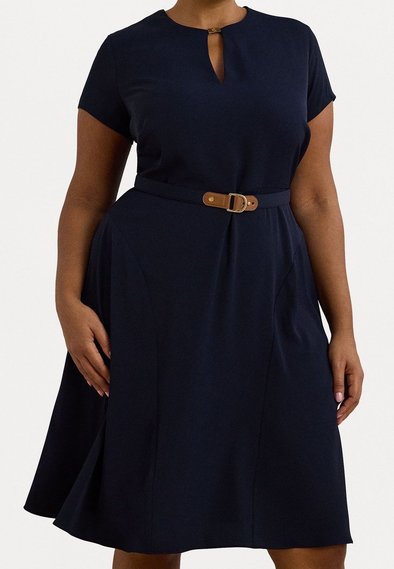 Frau trägt ein marineblaues knielanges Kleid mit kurzen Ärmeln, einem kleinen Schlüssel-Ausschnitt vorne und einem schmalen Gürtel mit einer goldenen Schnalle in der Taille.