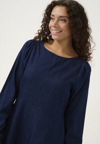 Marineblauwe denim jurk met een bootnek, lange pofmouwen en subtiele naaddetails. De stof heeft een gladde textuur en een lichte glans.