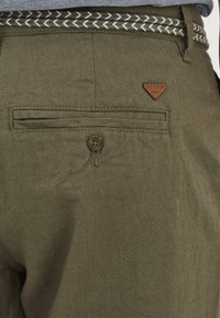 Pantalons vert olive avec une finition texturée, présentant une poche arrière avec un bouton et un petit patch en cuir marron. Ceinture tissée en blanc et noir.