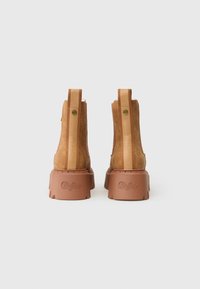 Par de botines de ante color beige con suelas de goma marrón gruesas y tiradores verticales embellecidos con texto en la parte trasera.