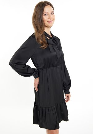 Musta satiini mekko, jossa on pitkät hihat, solmimiskaulus, kerääntynyt vyötärö ja levenevä röyhelöhelma. Sujuva rakenne ja polvipituinen malli.
