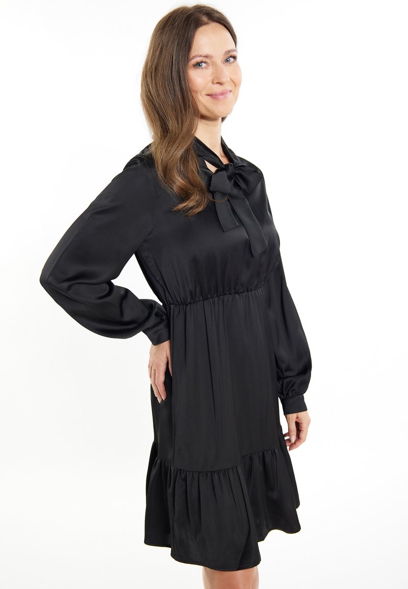 Abito in satin nero con maniche lunghe, colletto a fiocco, vita arricciata e orlo svasato con rouches. Tessuto liscio e design a lunghezza ginocchio.