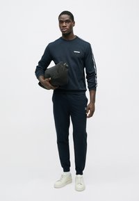 Muž oblečený v tmavomodrej tréningovej súprave a bielych teniskách drží čiernu športovú tašku, stojaci pred jednofarebným svetlým pozadím.