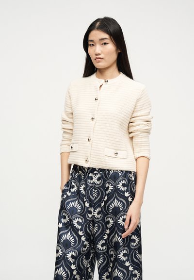 Ασιατική γυναίκα με μακριά σκούρα μαλλιά, φορώντας ένα βεραμάν υφασμένο ζακέτα με κουμπιά και ναυτικό παντελόνι με λευκά λουλουδάτα σχέδια.