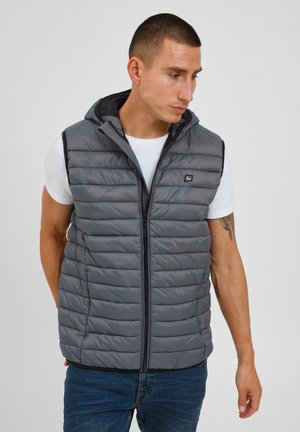 Mand med kort hår iført en grå quiltet, lynlåst, ærmeløs hættetrøje over en hvid t-shirt og blå jeans, kigger ned mod højre.