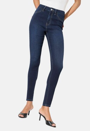 Femme portant un jean skinny bleu foncé taille haute et des sandales noires à talons hauts et bouts ouverts, debout les mains derrière le dos.