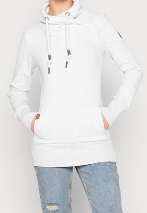 Person iført en lysegrå hættetrøje med snore og hænder i frontlommerne, kombineret med revnede lyseblå jeans.