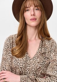 Kobieta o długich jasnobrązowych włosach, ubrana w brązowy kapelusz z szerokim rondem i bluzkę w panterkę z marszczonymi detalami, ze skrzyżowanymi ramionami.