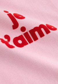 Světlo rožnata tkanina ima rdeča, žametna črka z izrazom "je t'aime" v krepki, igrivi pisavi. Gladek tekstura s subtilnim sijajem.