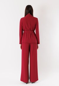 Completo rosso su misura con vita stretta e pantaloni a gamba larga. Tessuto liscio con un design strutturato e rever a punte. Vista posteriore mostrata.