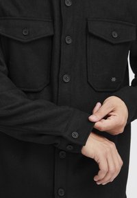 Black button-up shirt with two chest pockets, made of soft fabric. Features black buttons and cuff details. Clean, minimal design.