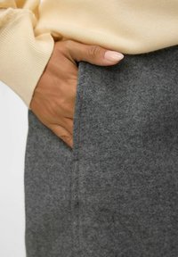 Jupe en feutre gris avec une poche latérale, montrant un gros plan d'une main reposant contre le tissu. Texture douce, design structuré.